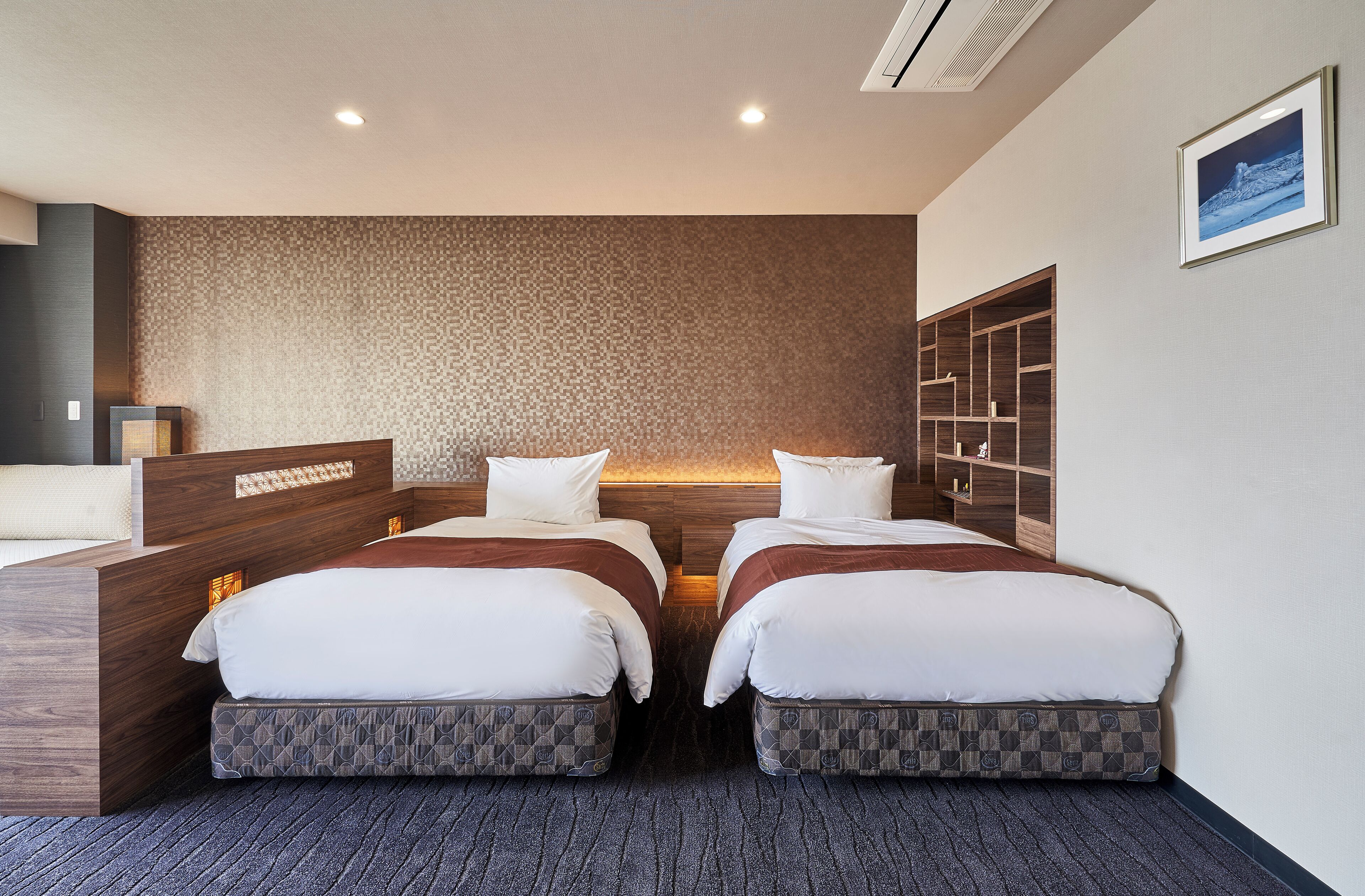 A hotel room with two beds, a wooden headboard, a patterned wall, and a built-in shelving unit.