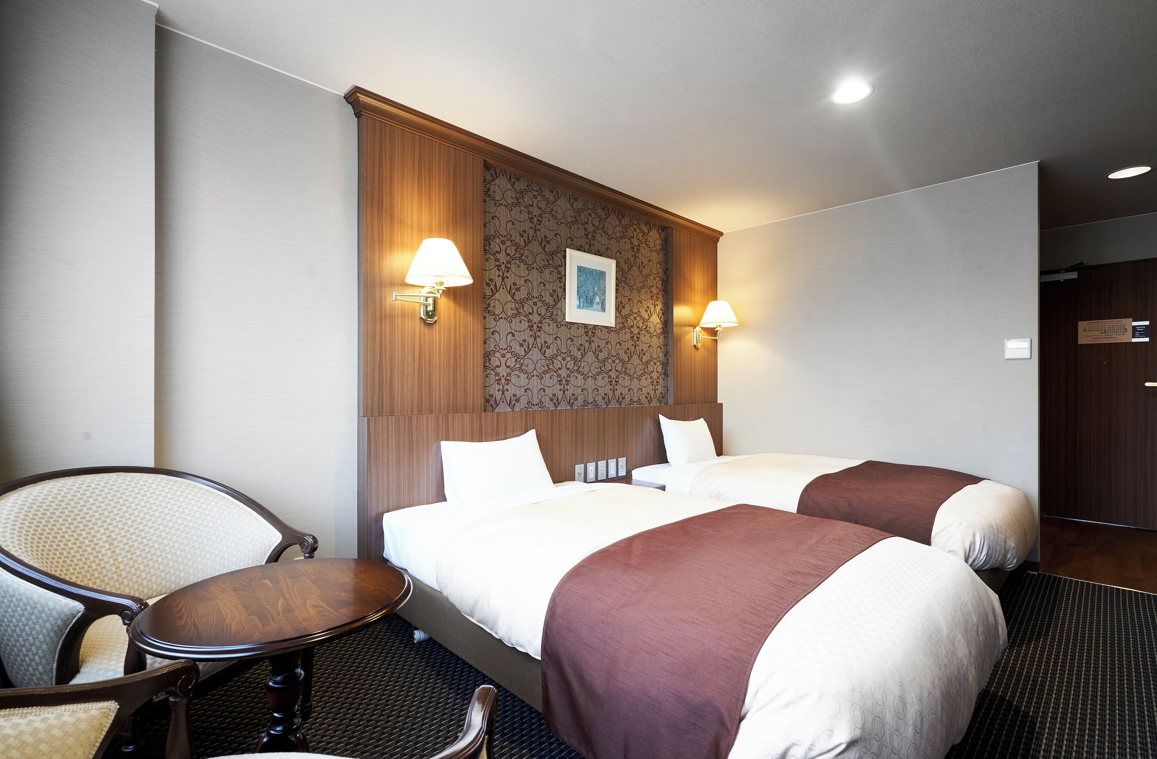 Ein Hotelzimmer mit zwei Betten, einem Sessel, einem kleinen Tisch und Wandlampen.