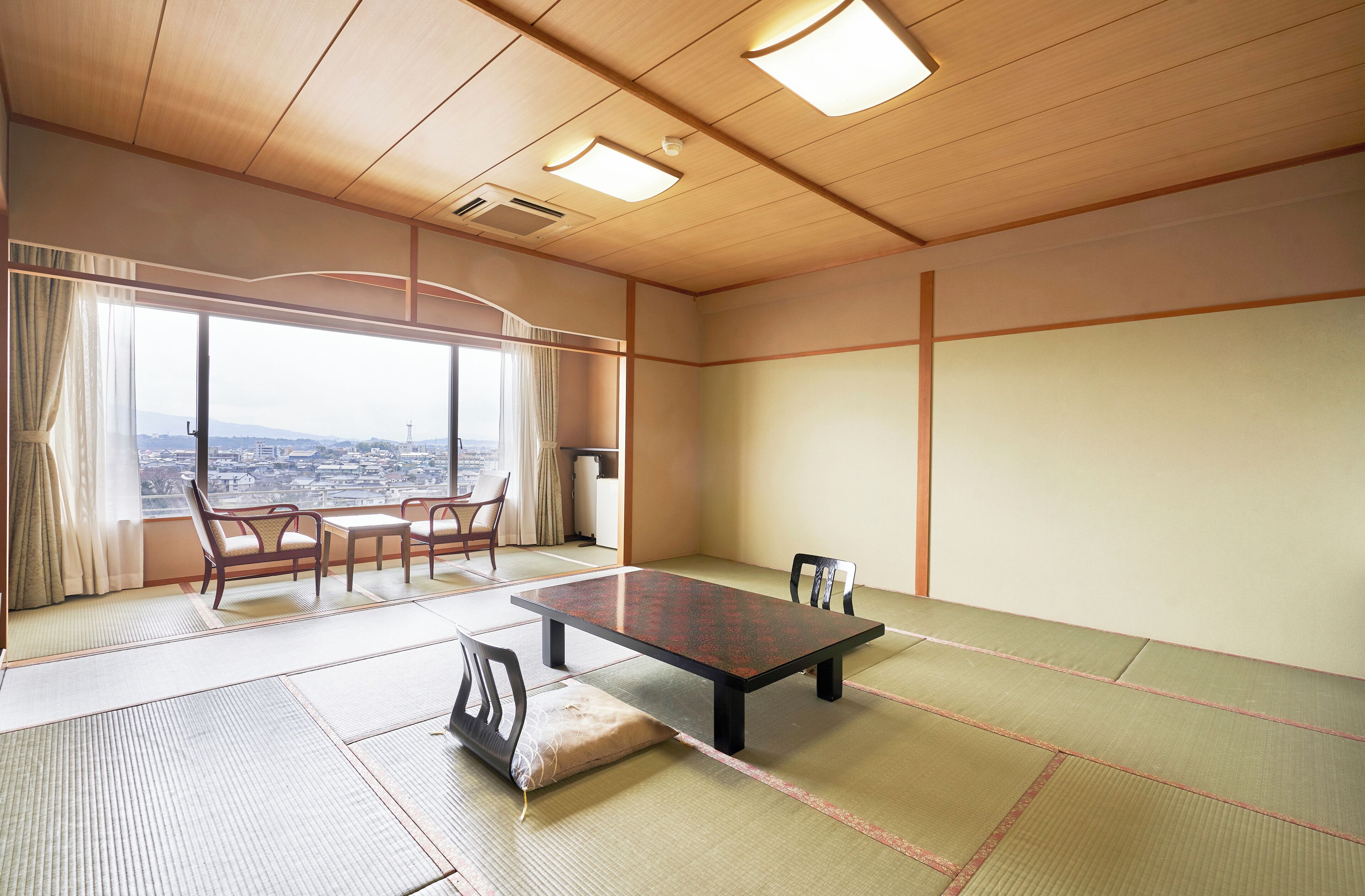 Ein Raum mit Tatami-Matten, einem niedrigen Tisch, Stühlen, großen Fenstern mit Blick auf die Stadt und einer Holzbodenverkleidung.