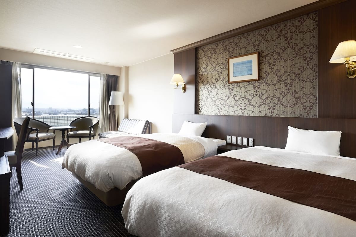 Een hotelkamer met twee bedden, een bureau, een stoel en uitzicht op de stad.