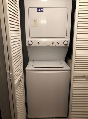 Une petite buanderie blanche, avec une pancarte indiquant « NOUVEAU », un lave-linge à chargement frontal et un sèche-linge.