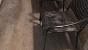 Eine Reihe Metallstühle mit geflochtenen Sitzflächen und Rückenlehnen, auf einem Betonboden neben einer beigen Tür und einem Fenster mit Insektenschutzgitter.