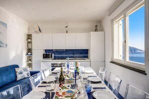 Un comedor con una mesa preparada para cuatro personas, un sofá azul, armarios blancos y vistas al mar.