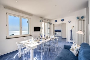 Un coin repas avec une table dressée pour six personnes, des chaises transparentes, un canapé bleu et un téléviseur fixé au mur.