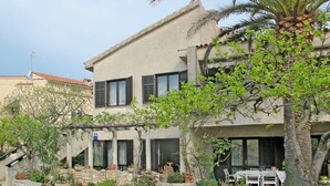 Ein zweigeschossiges Haus mit einer Steintreppe, die zum Eingang führt, umgeben von verschiedenen Kübelpflanzen und einem Palmenbaum.