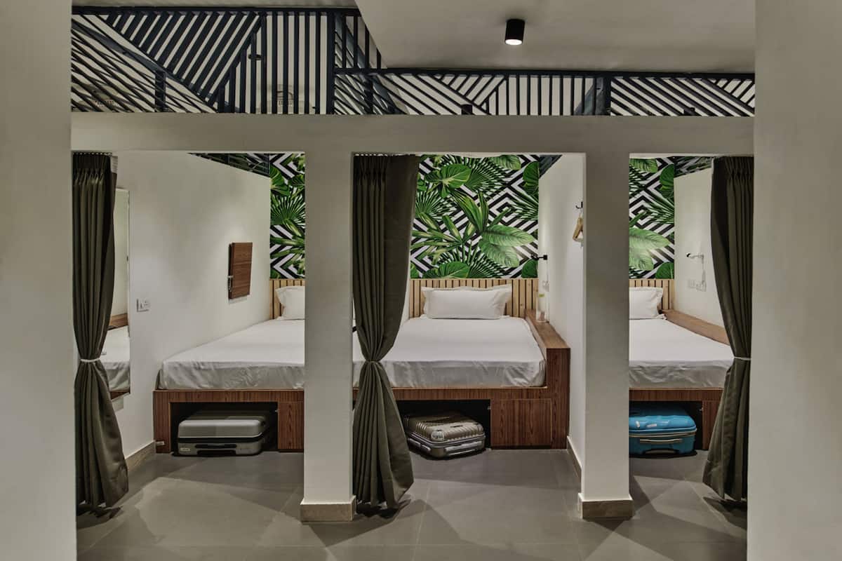 Une chambre d’hôtel avec trois lits, chacun doté d’une tête de lit en bois, de draps blancs et d’une valise placée sous chaque lit. La chambre présente un design moderne, avec un mur orné d’un motif géométrique derrière les lits et un escalier à rayures noires et blanches.