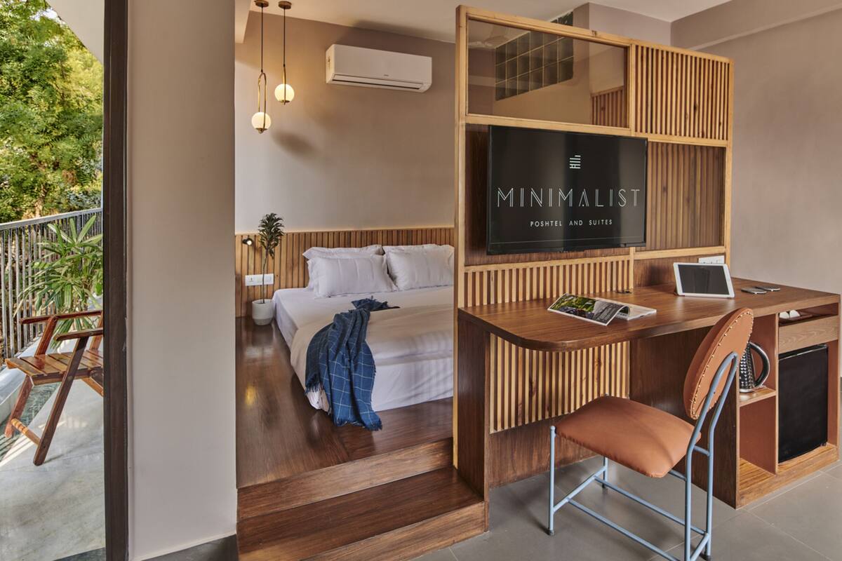 Une chambre d’hôtel moderne avec un bureau en bois, un ordinateur portable et un fauteuil confortable. On y trouve un lit avec du linge de maison blanc et une couverture bleue. La chambre dispose d’une grande fenêtre et offre une vue sur la verdure environnante.