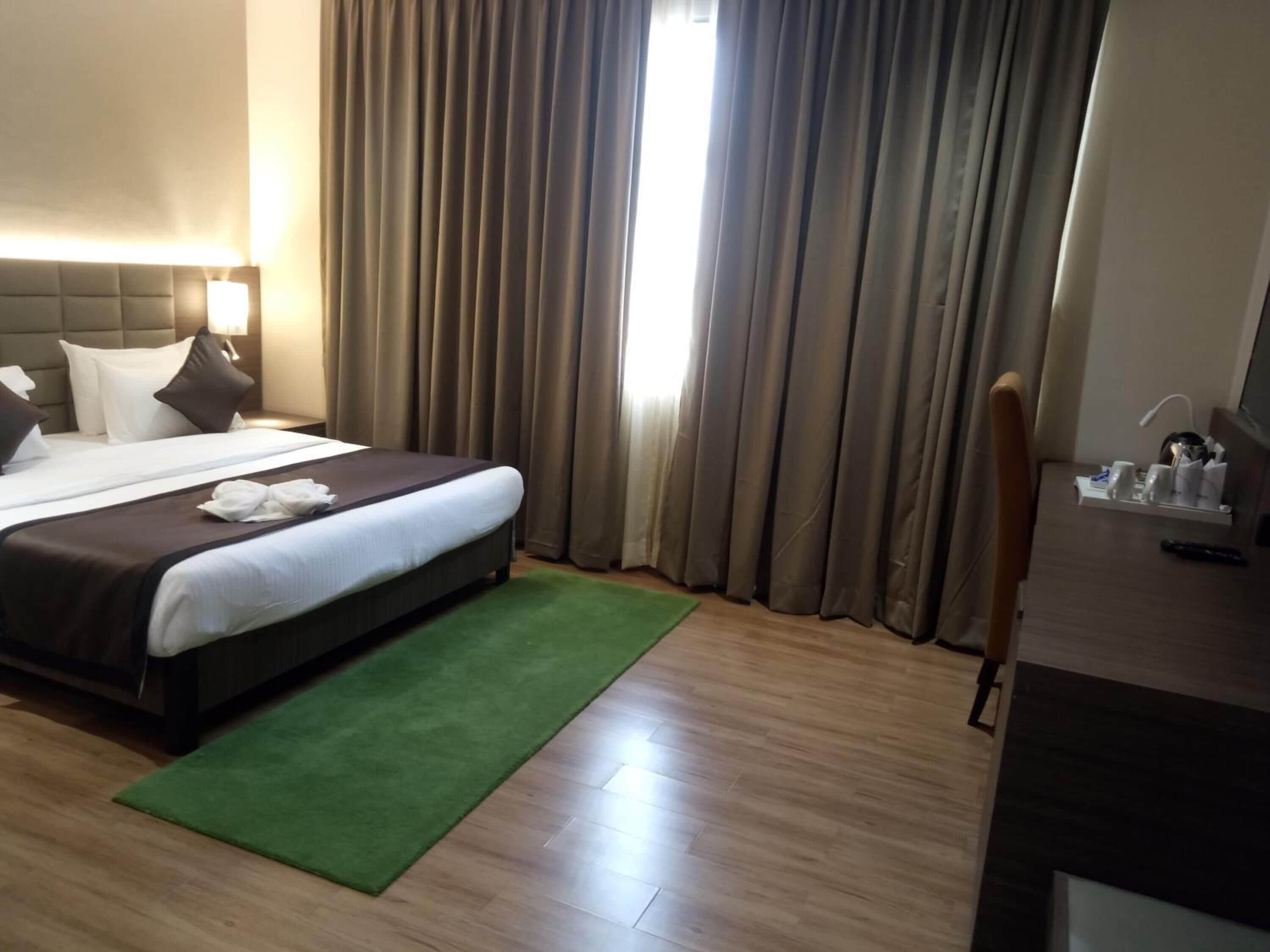 A hotel room with a bed, a desk, a chair, a green rug, and a window with curtains.