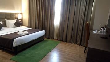 A hotel room with a bed, a desk, a chair, a green rug, and a window with curtains.