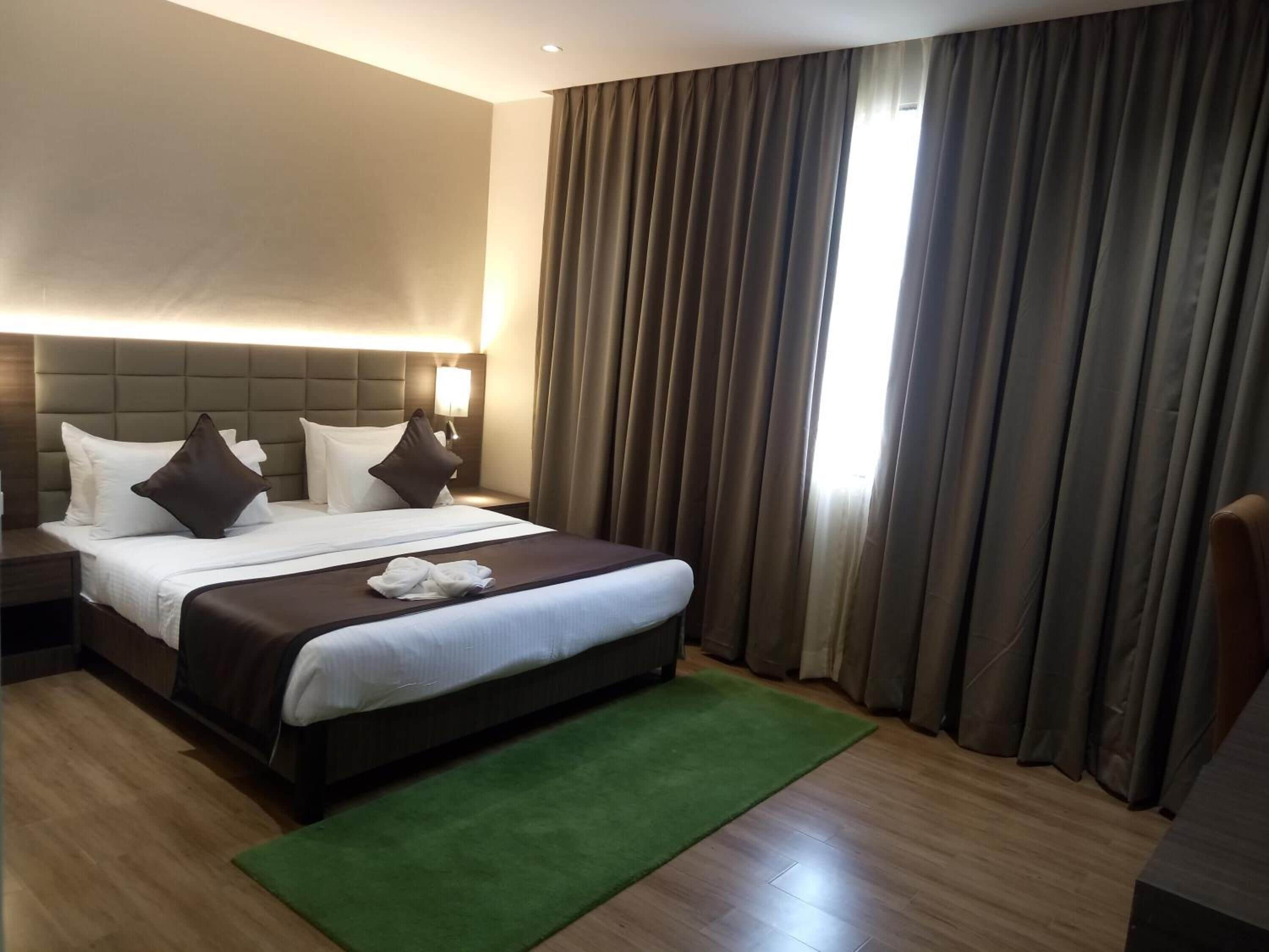 A hotel room with a large bed, a green rug, a headboard with built-in lighting, and a view of the outdoors through curtains.