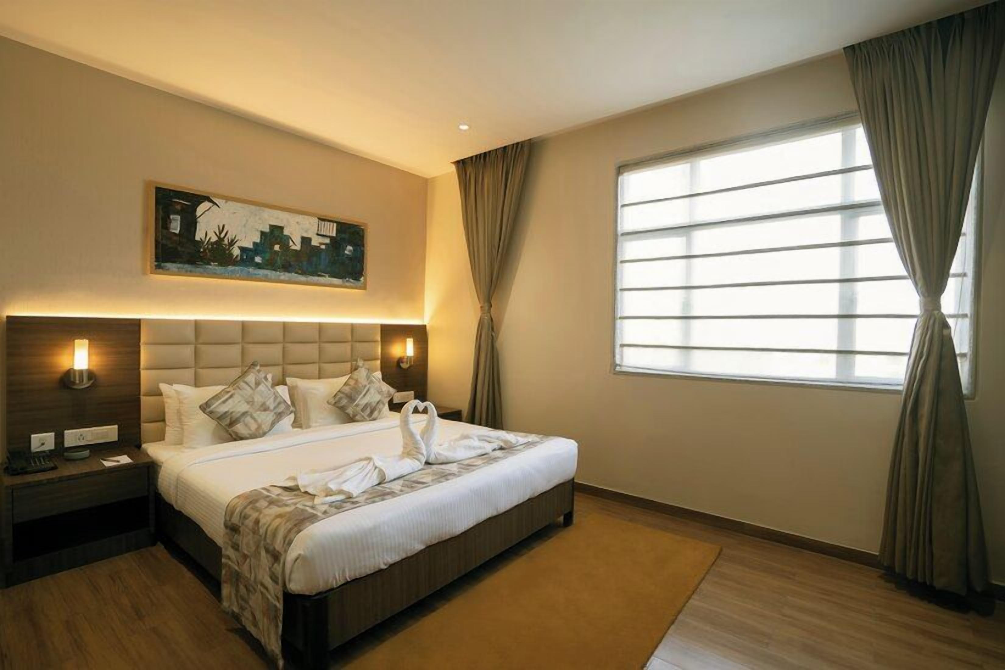 A hotel room with a large bed, a nightstand, a wall-mounted lamp, a window with curtains, and a painting on the wall.