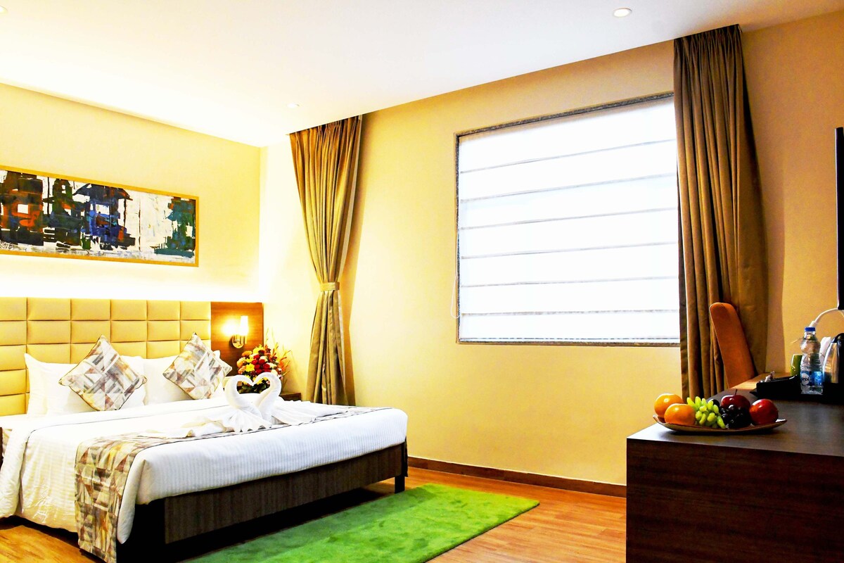 A hotel room with a bed, a green rug, a fruit bowl, and a painting on the wall.
