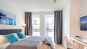 Ein modernes Hotelzimmer mit Bett, Schreibtisch und Stuhl. Zwei Fenster mit Vorhängen, ein Bild an der Wand und eine Lampe auf dem Nachttisch.