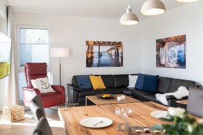 Un salon moderne avec un canapé en cuir noir, un fauteuil rouge et une table basse en bois. Deux tableaux sont accrochés au mur et représentent une jetée et un bateau.