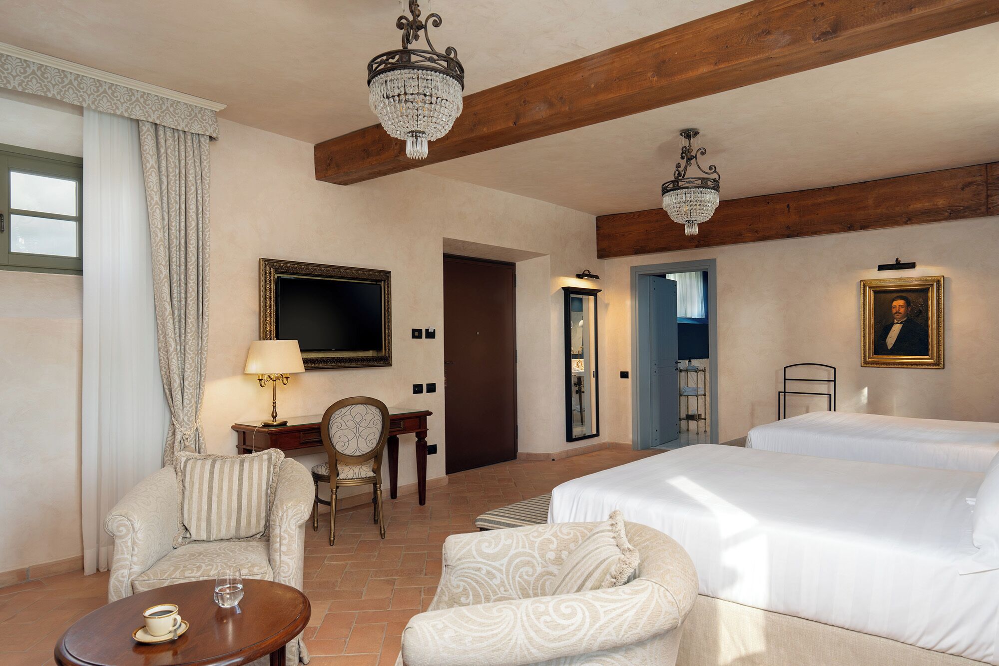 Habitación de hotel con cama, dos sillones, una mesa, televisor, cuadro y dos candelabros.