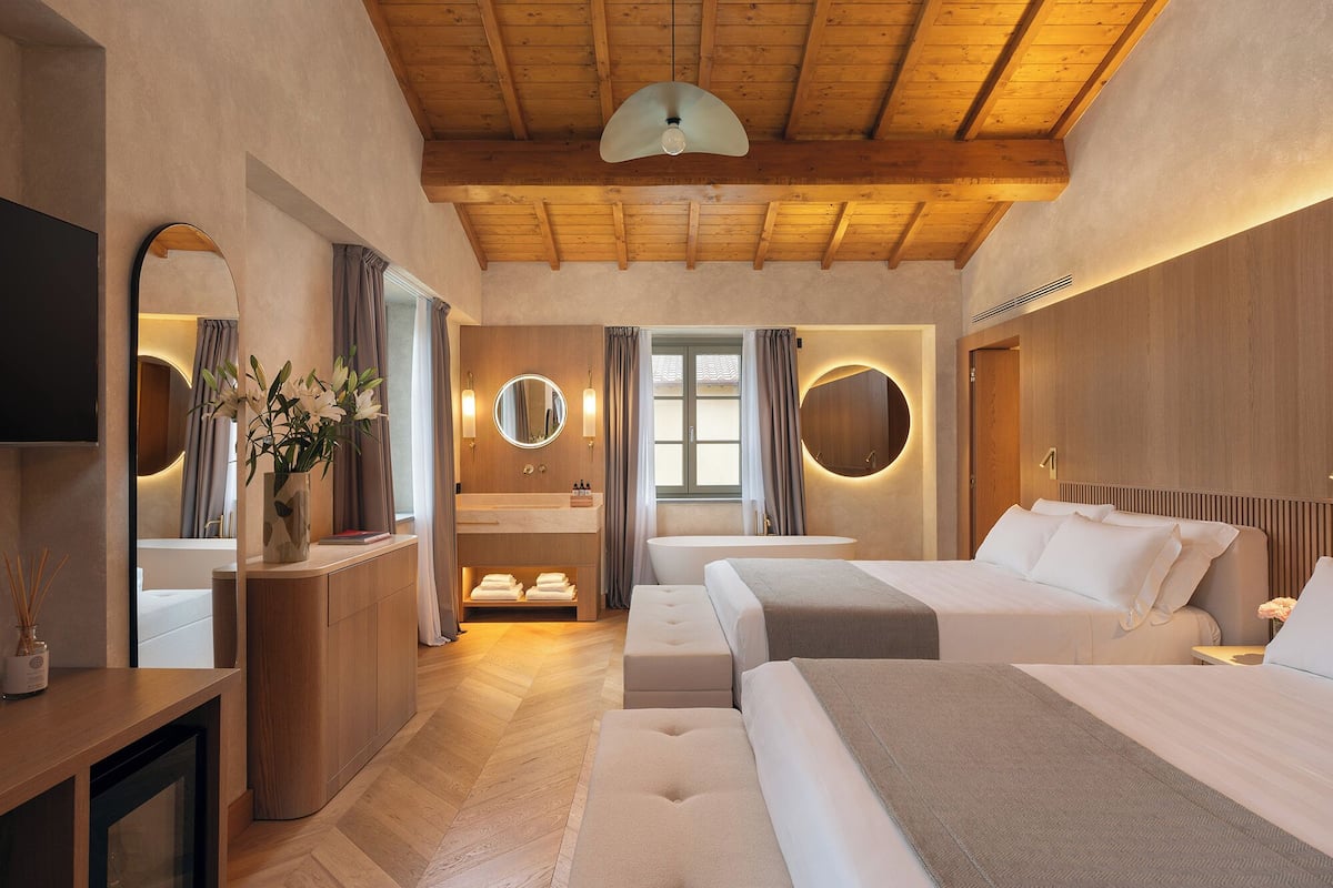 Ein modernes Hotelzimmer mit Holzboden, einem großen Bett, einem kleineren Bett, einem Frisiertisch mit Spiegel und einer Badewanne.