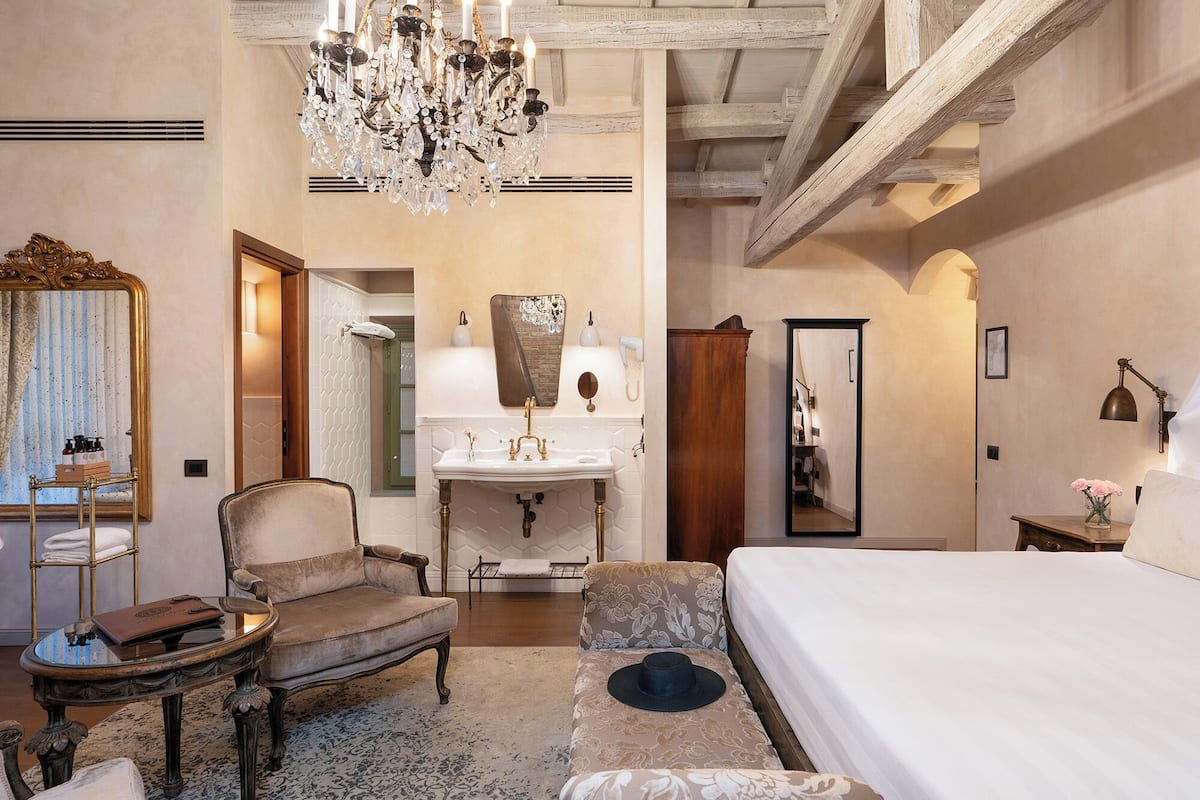 Ein Hotelzimmer mit einem großen Bett, einer Sitzecke mit Sessel und Tisch, einem Schminktisch mit Spiegel und Waschbecken sowie einer Kandelaber.