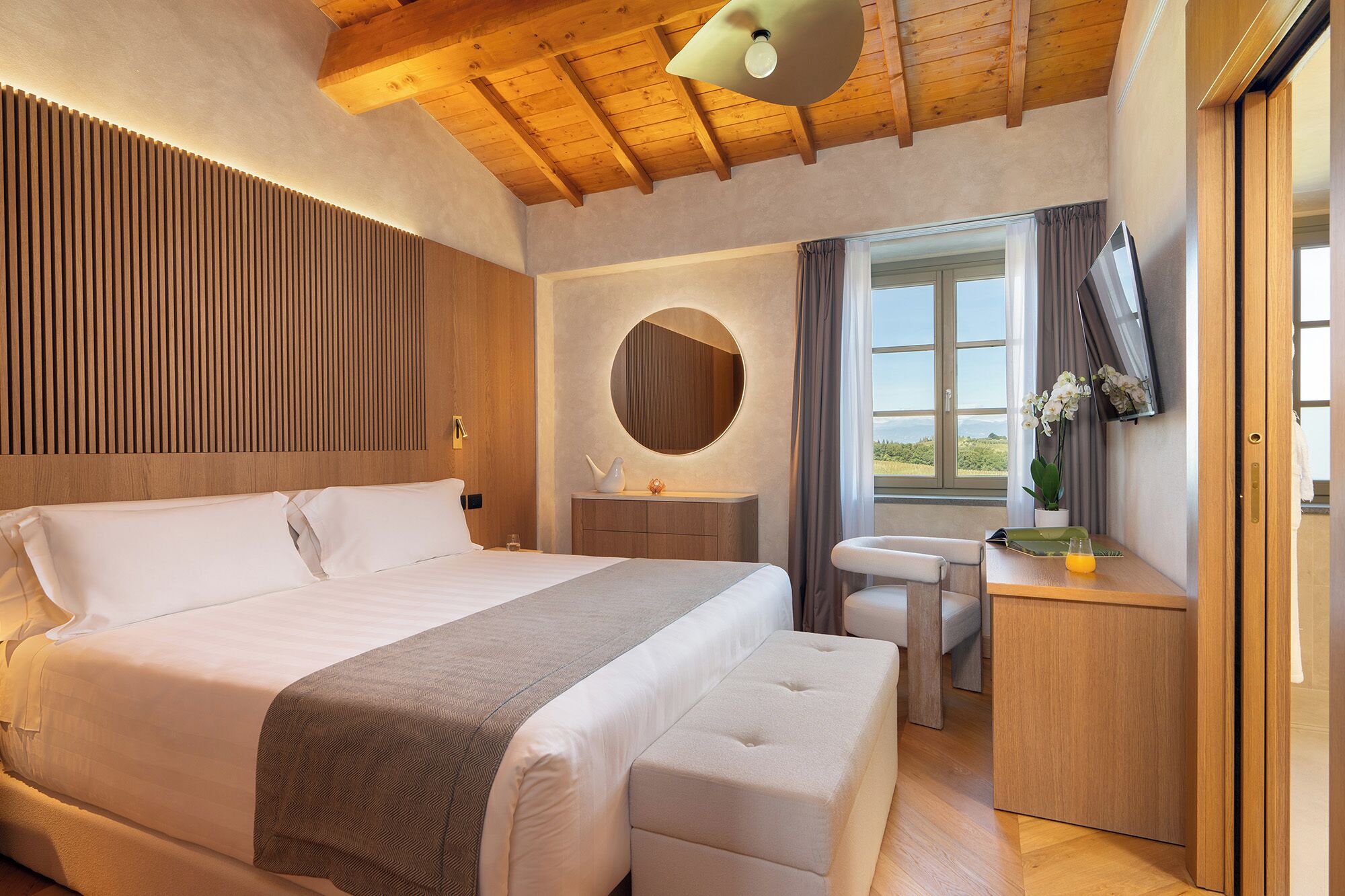 Une chambre avec un grand lit, une table de chevet, un miroir, une fenêtre donnant sur un beau paysage et un téléviseur.
