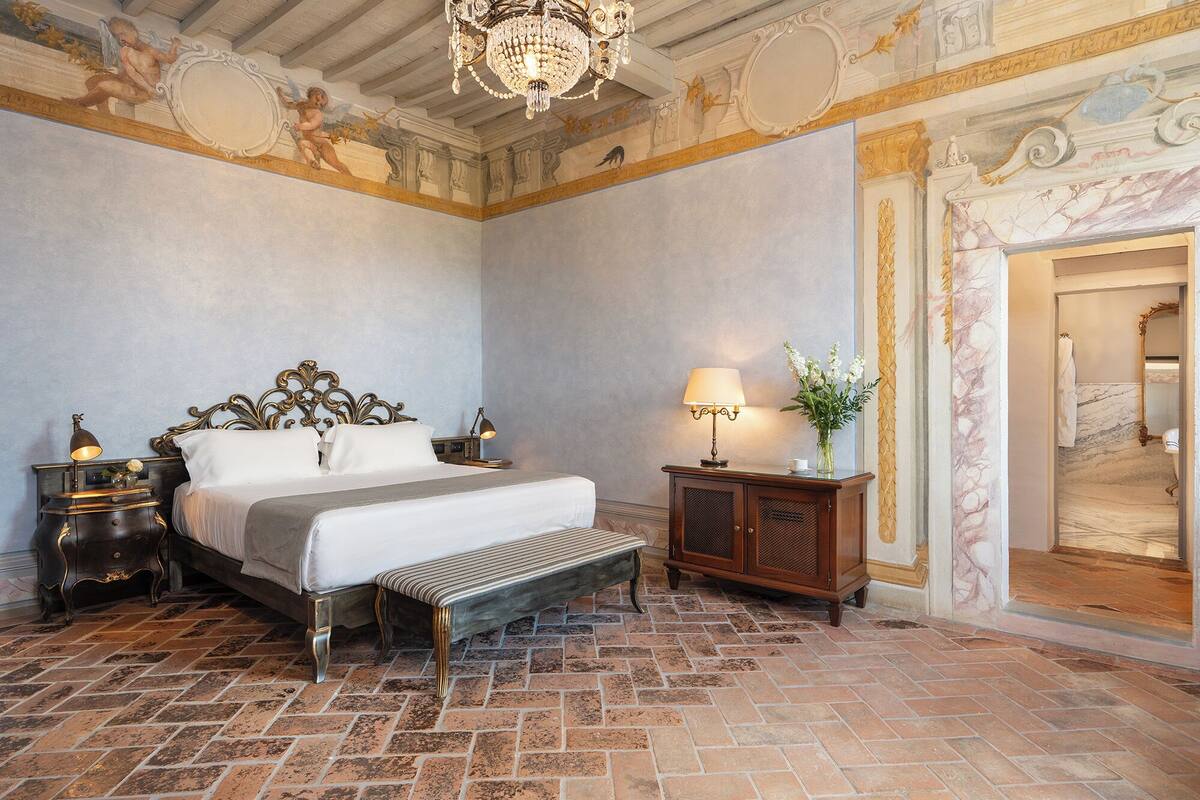 Ein Schlafzimmer mit einem großen Bett, einer Bank, einem Nachttisch, einer Lampe und einem Spiegel.