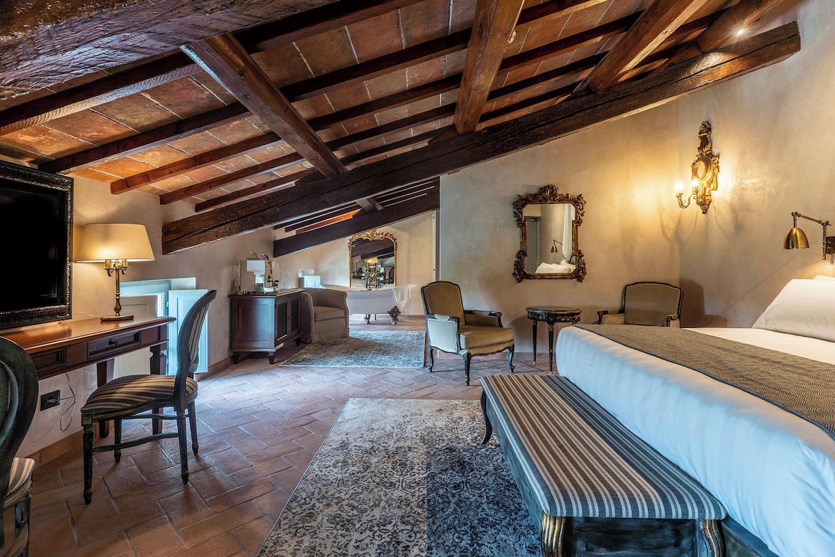 Ein Hotelzimmer mit Holzboden, einem Bett, einem Schreibtisch, einem Stuhl, einem Spiegel und einem im Hintergrund sichtbaren Badezimmer.