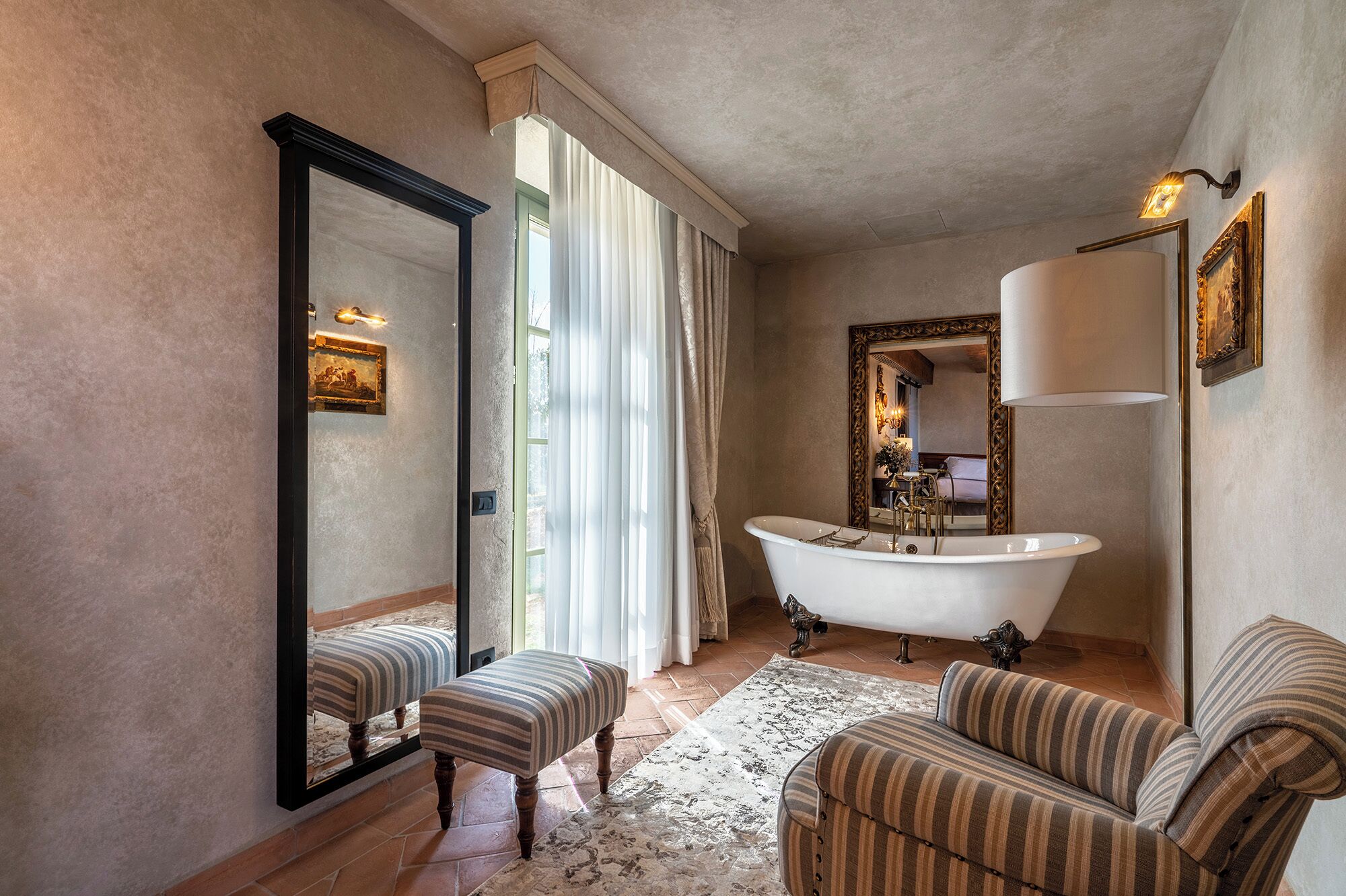 Un baño con una bañera independiente, un espejo grande y sillones tapizados a rayas.