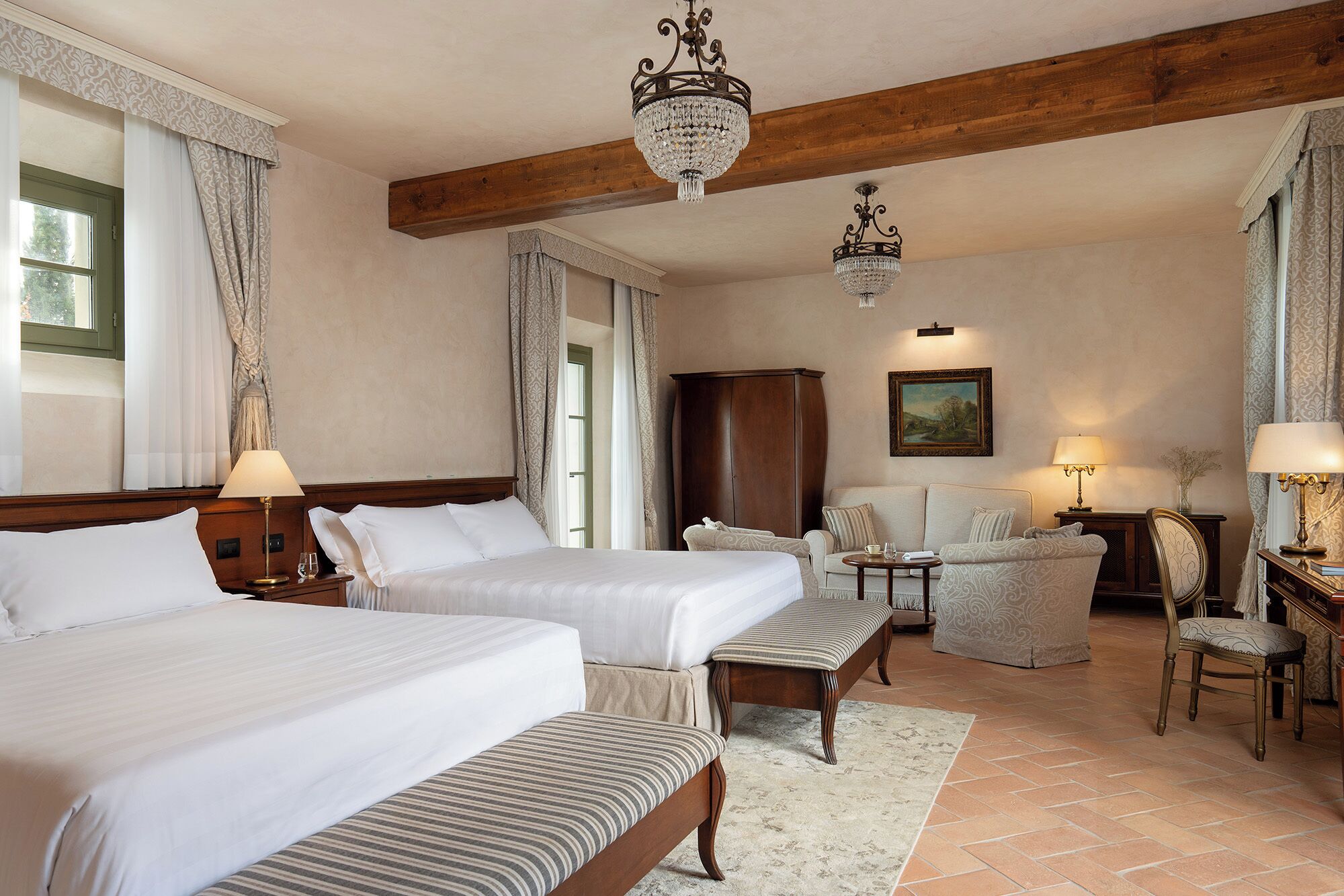 A hotel room with two beds, a wooden ceiling, chandeliers, a sofa, a chair, a cabinet, and a painting on the wall.