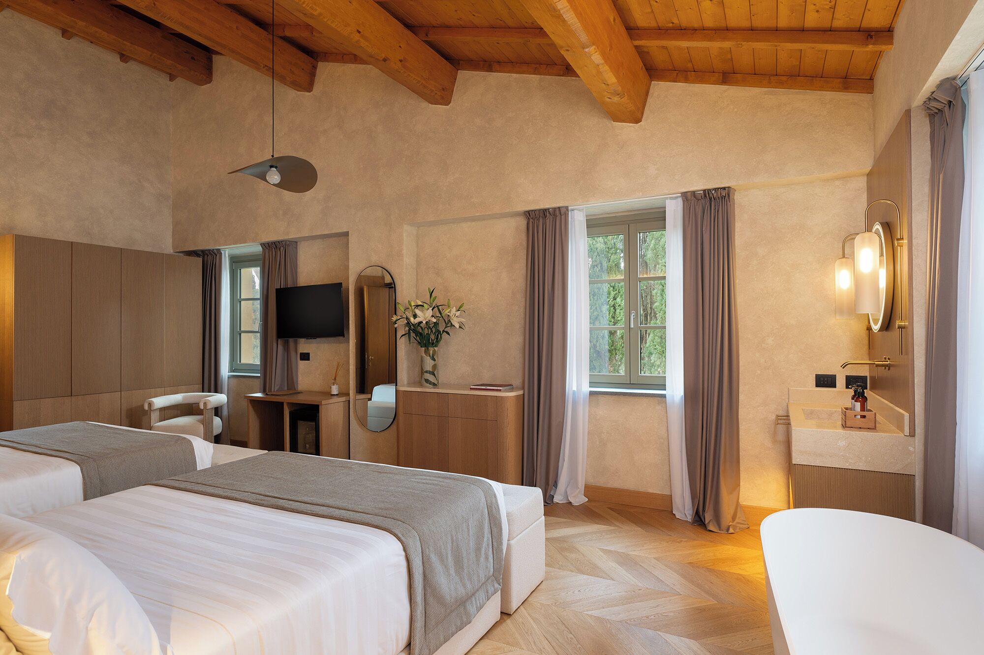 Une chambre d’hôtel avec deux lits, une télévision, un miroir, une coiffeuse avec un lavabo et une fenêtre avec des rideaux.