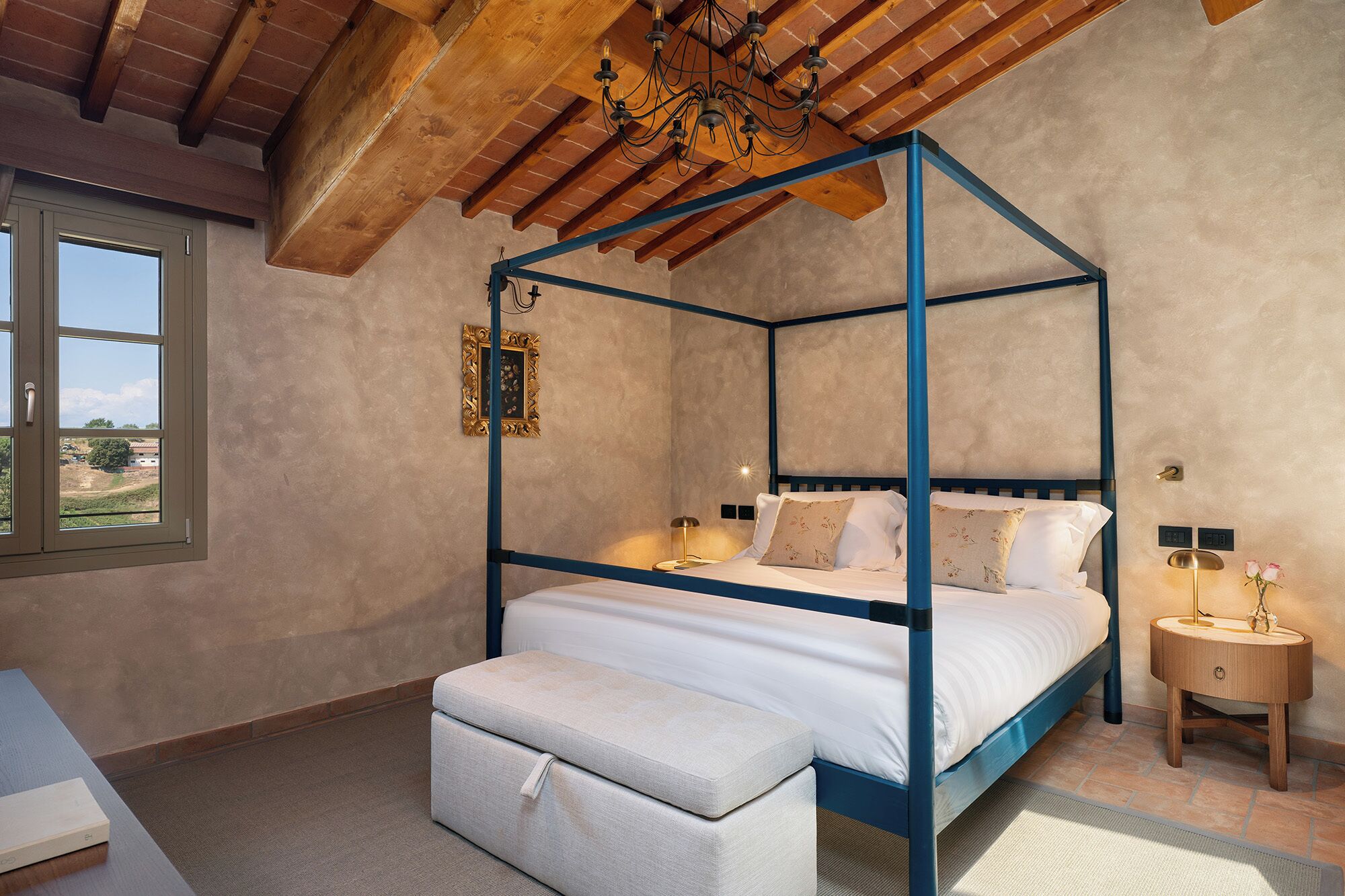 A four-poster bed with a bench, a bedside table, a nightstand, and a window with a view.