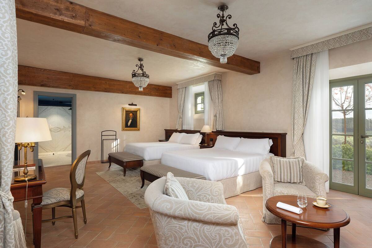 Ein Hotelzimmer mit zwei Betten, einer Sitzecke mit Sessel und Tisch, einer Lampe, einem Bild an der Wand und einer Chandelieber.