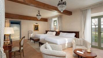 Quarto de hotel com duas camas, área de estar com uma poltrona e mesa, abajur, quadro na parede e lustre.