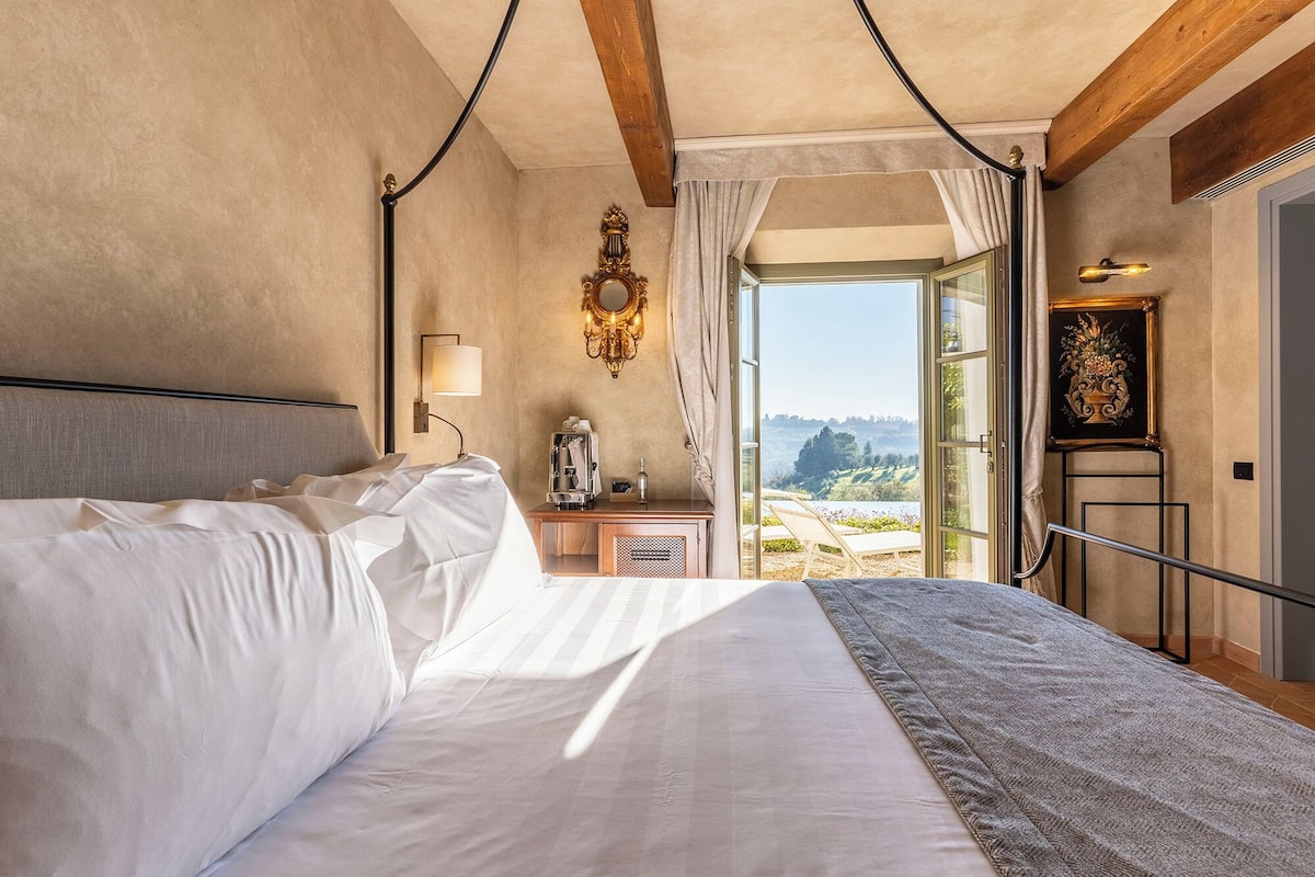 Ein Hotelzimmer mit einem großen Bett, einem Nachttisch mit Lampe, einem Spiegel und Blick ins Freie.