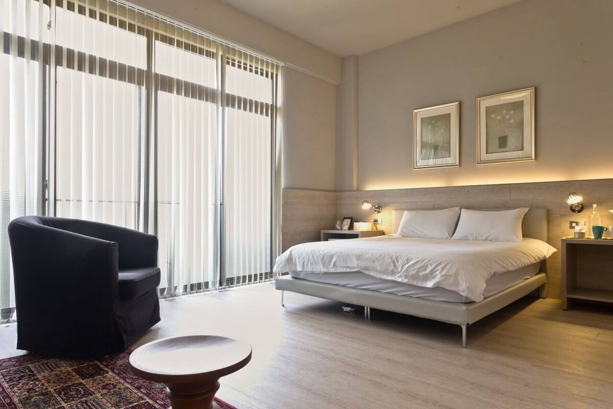 Quarto de hotel moderno com uma cama grande, uma poltrona preta, uma mesa redonda pequena e vista através de janelas do chão ao teto.