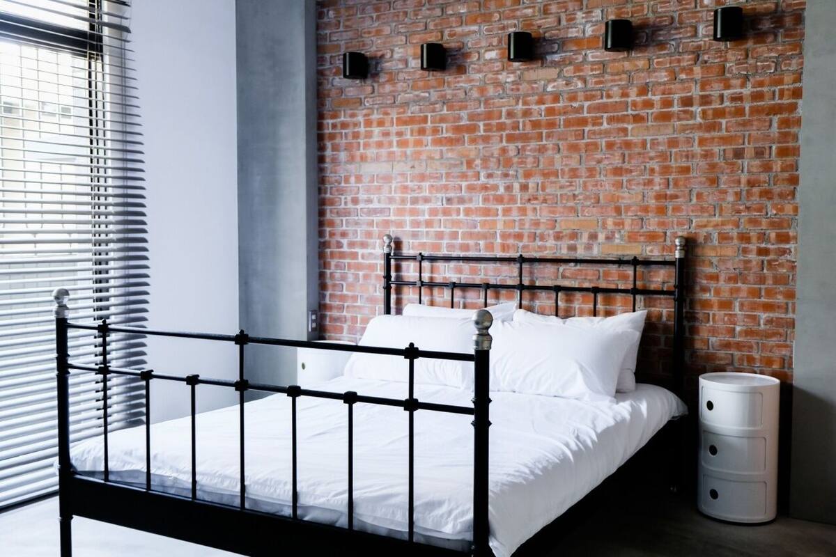 Quarto moderno com parede de tijolos, estrutura de cama em metal, roupa de cama branca e janela com persianas.