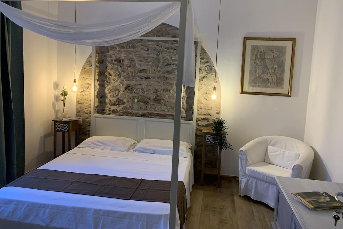 Quarto com parede de pedra, cama com dossel, cadeira branca e uma pequena mesa com abajur.