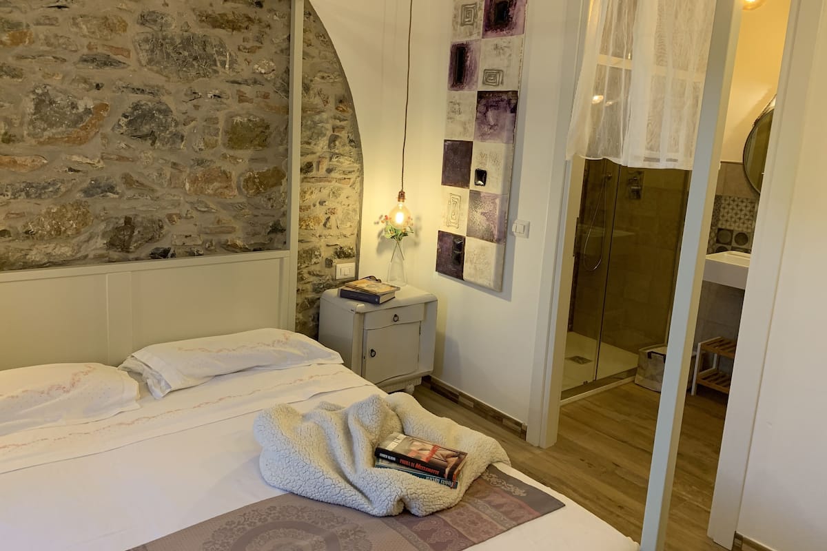 Quarto com parede de pedra, mesa de cabeceira com livros, cama com manta e área de chuveiro.