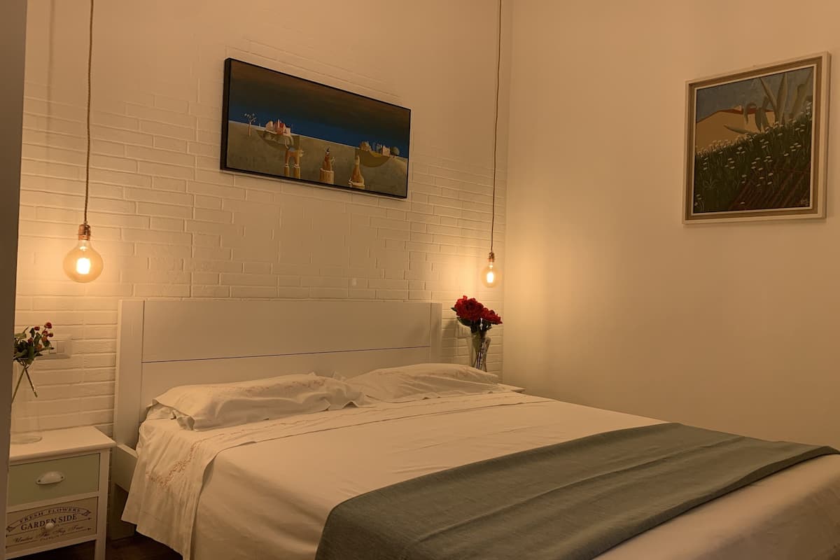 Cama bem arrumada com lençóis brancos, manta cinza e um vaso com flores vermelhas em uma mesinha de cabeceira. Duas lâmpadas pendem do teto, e um quadro é visível na parede acima da cama.