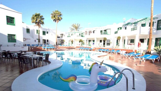 Zona de piscina con sillones de descanso, un flotador con forma de unicornio y un edificio de paredes blancas y ventanas verdes.