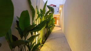 A narrow pathway flanked by lush green plants and a white wall, leading to a building with a red-tiled roof. - Danka Villa (Denpasar)