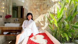 A person sitting in a bathtub filled with red and white rose petals, with a stone wall and a large plant in the background. - Danka Villa (Denpasar)