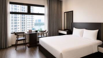 A hotel room with a large bed, a desk with two chairs, a view of buildings through the window, and a mirror on the wall.