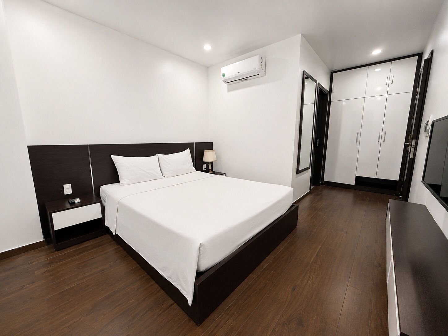 Una camera d'albergo moderna con un letto grande, un comodino, una lampada, un condizionatore d'aria, un armadio e una televisione.