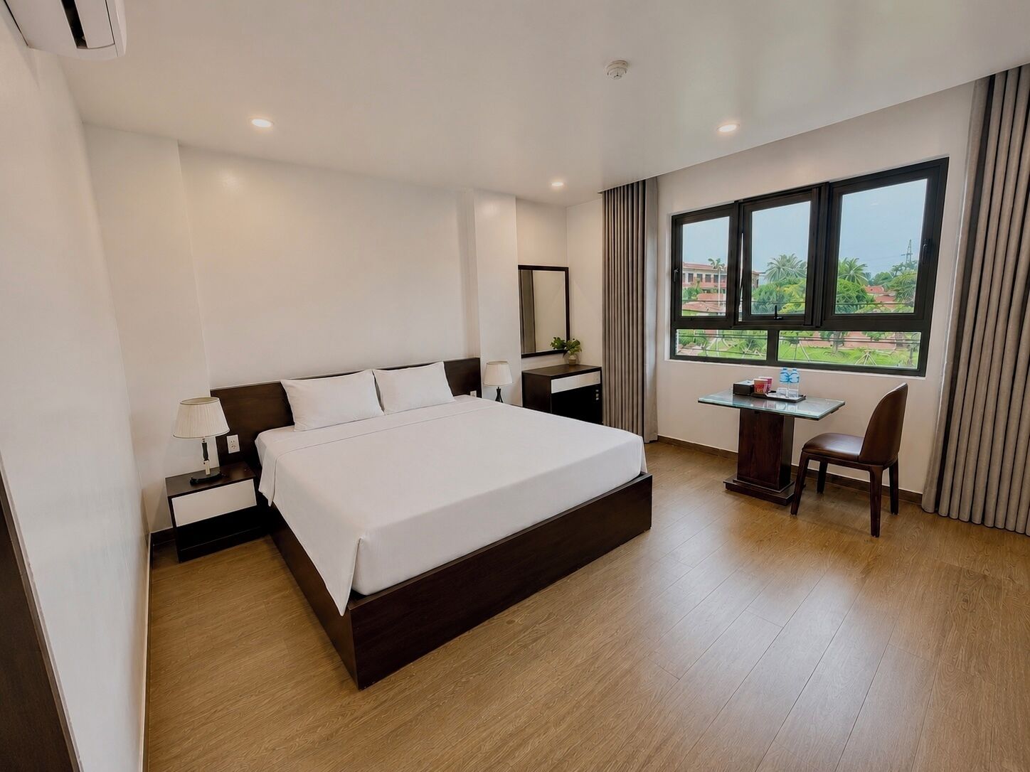 A hotel room with a large bed, a desk with a chair, a window with a view, and a nightstand with a lamp.