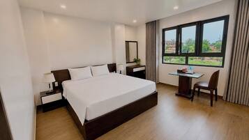 A hotel room with a large bed, a desk with a chair, a window with a view, and a nightstand with a lamp.