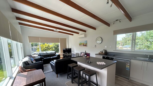 A modern kitchen with a central island, a dining area with a table and chairs, a living room with a sofa and a TV, and large windows offering a view of the outdoors.