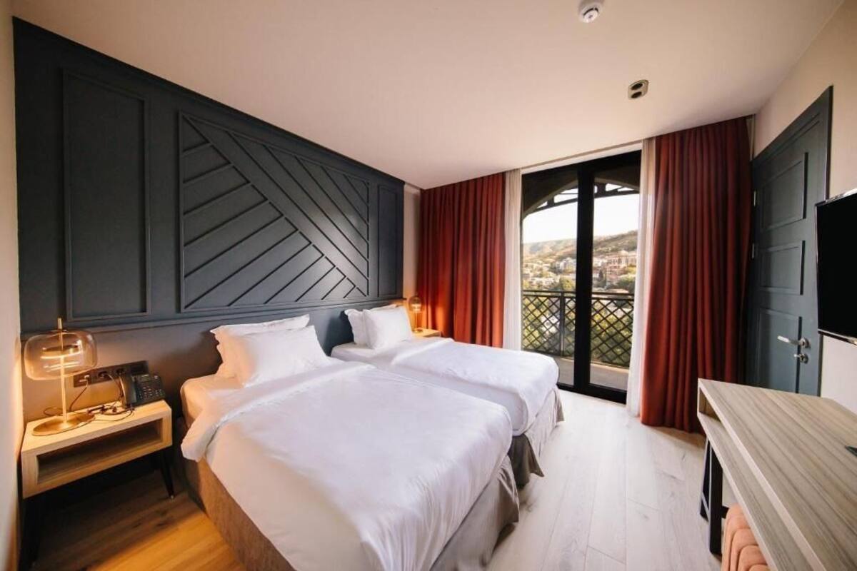 A hotel room with two beds, a desk, a TV, and a balcony with red curtains.