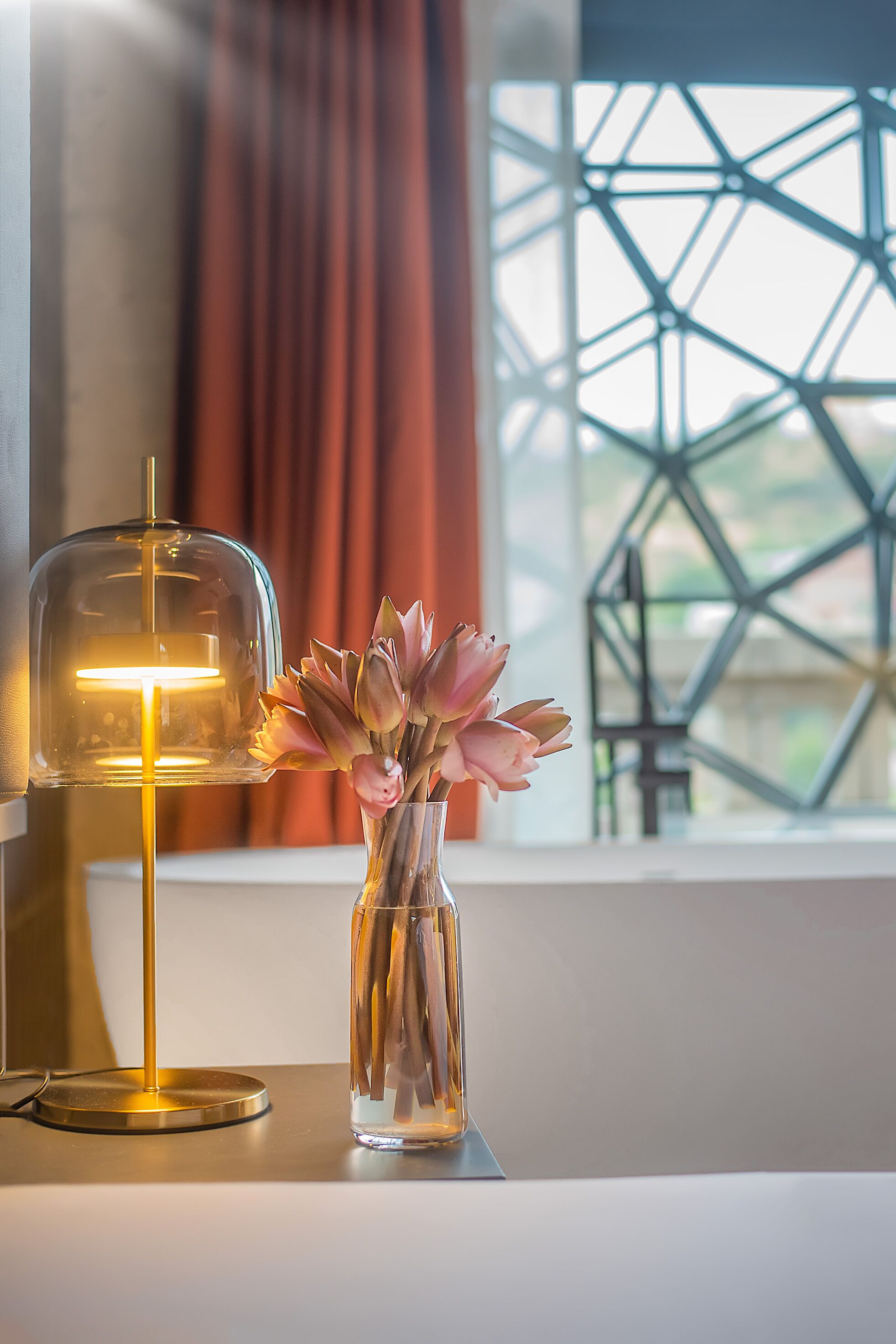 A clear glass vase with pink flowers on a table next to a lamp with a clear glass shade and a gold base.