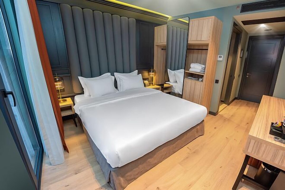 A modern hotel room with a large bed, bedside tables, a wooden wardrobe, and a desk.