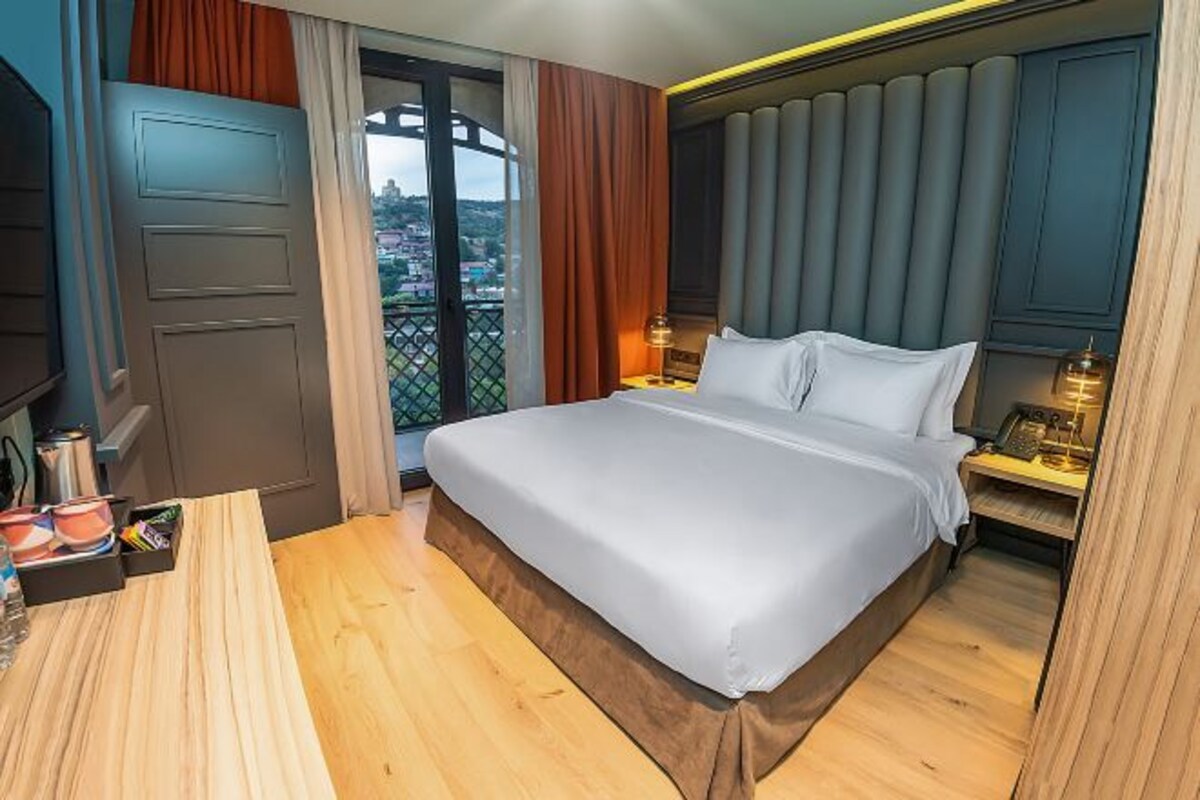 A hotel room with a large bed, a desk with a chair, a window with curtains, and a view of a cityscape.