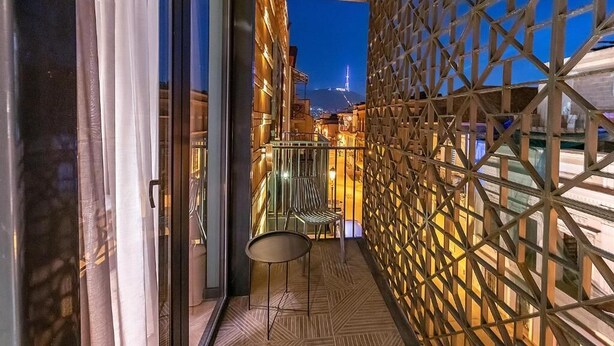 Un balcón con vista a un paisaje urbano al atardecer, que presenta una celosía geométrica y una pequeña mesa con sillas.
