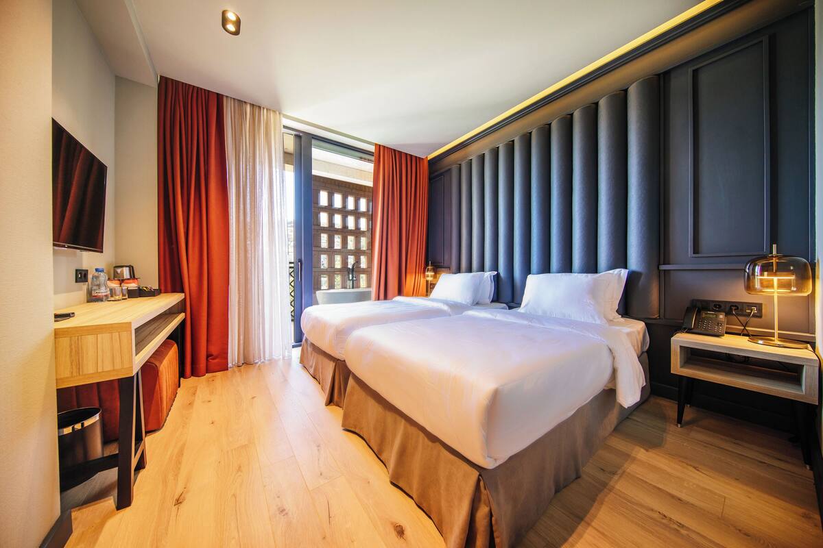 A hotel room with a large bed, a desk with a chair, a TV, and a bedside table with a lamp.